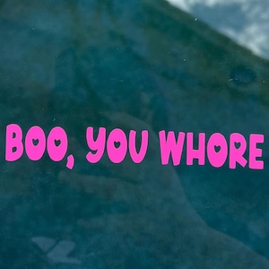 Op de afbeelding: Roze tekst op een blauwe achtergrond met de tekst "Boo, You Whore". De tekst is in een vet, afgerond lettertype, met hartvormige symbolen ter vervanging van de letter "O".