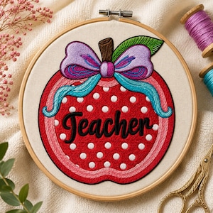 May include: A circular wooden embroidery hoop containing a fabric piece with a stitched red apple design. The apple features white polka dots, a brown stem, a green leaf, and a large purple and teal bow. The word Teacher is stitched in black.