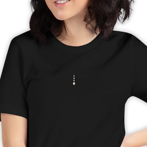 以下が含まれることがあります： 胸に小さな垂直のオフホワイトのデザインが施された黒いTシャツ。デザインは、4つの小さな、等間隔に配置されたドットで構成されており、一番下のドットはわずかに大きく、涙の形をしています。