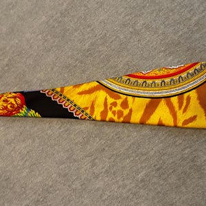 May include: A patterned silk scarf with a vibrant design. The scarf features a tiger stripe pattern in shades of yellow and brown, with ornate borders in red, gold, and black. A detailed illustration of a face is visible on the black section.