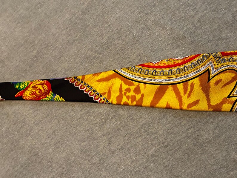 May include: A patterned silk scarf with a vibrant design. The scarf features a tiger stripe pattern in shades of yellow and brown, with ornate borders in red, gold, and black. A detailed illustration of a face is visible on the black section.