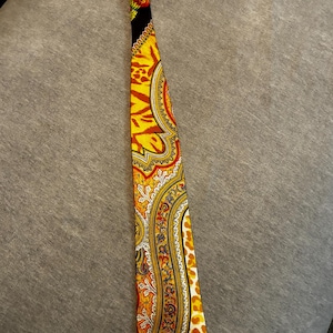 May include: A colorful necktie with a vibrant paisley pattern in shades of yellow, orange, and red. The tie features a black top section with a floral design and a leopard print on the edge. The tie is made of a silky material.