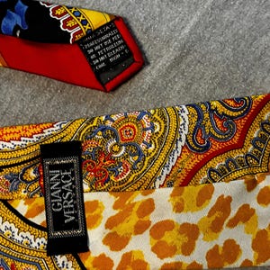 May include: Close-up of two silk neckties. One tie features a red, yellow, and black paisley design. The other tie has a yellow and orange paisley pattern with a leopard print lining. The label reads "Gianni Versace".