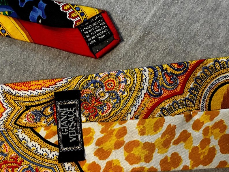 May include: Close-up of two silk neckties. One tie features a red, yellow, and black paisley design. The other tie has a yellow and orange paisley pattern with a leopard print lining. The label reads "Gianni Versace".