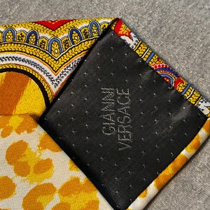 May include: Close-up of a patterned silk tie with a black backing. The tie features yellow, orange, red, and blue designs. The black backing has the text "GIANNI VERSACE" in white.