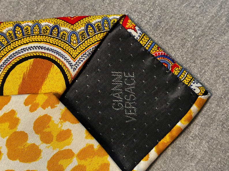 May include: Close-up of a patterned silk tie with a black backing. The tie features yellow, orange, red, and blue designs. The black backing has the text "GIANNI VERSACE" in white.
