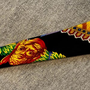 May include: A black silk scarf with a detailed design. The scarf features a yellow and red face with a red head covering, surrounded by green and blue accents. The border has a yellow and red pattern.