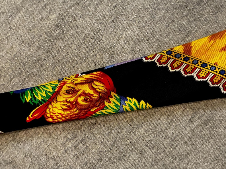 May include: A black silk scarf with a detailed design. The scarf features a yellow and red face with a red head covering, surrounded by green and blue accents. The border has a yellow and red pattern.