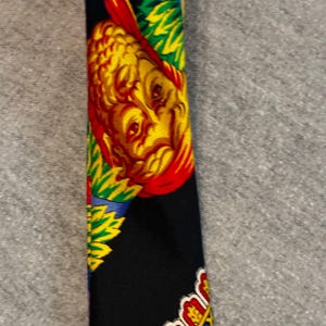 May include: A black necktie with a colorful pattern. The design features a yellow and red face, with green, blue, and yellow accents. The tie's bottom edge has a decorative border with red, yellow, and blue details.