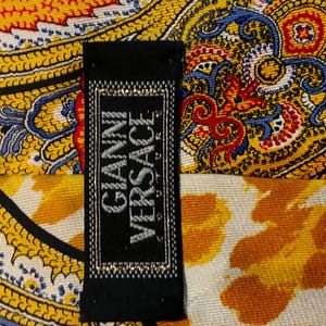 May include: A black label with the text "GIANNI VERSACE" in silver lettering is displayed against a backdrop of a colorful patterned fabric. The fabric features a paisley design with yellow, orange, blue, and red hues.