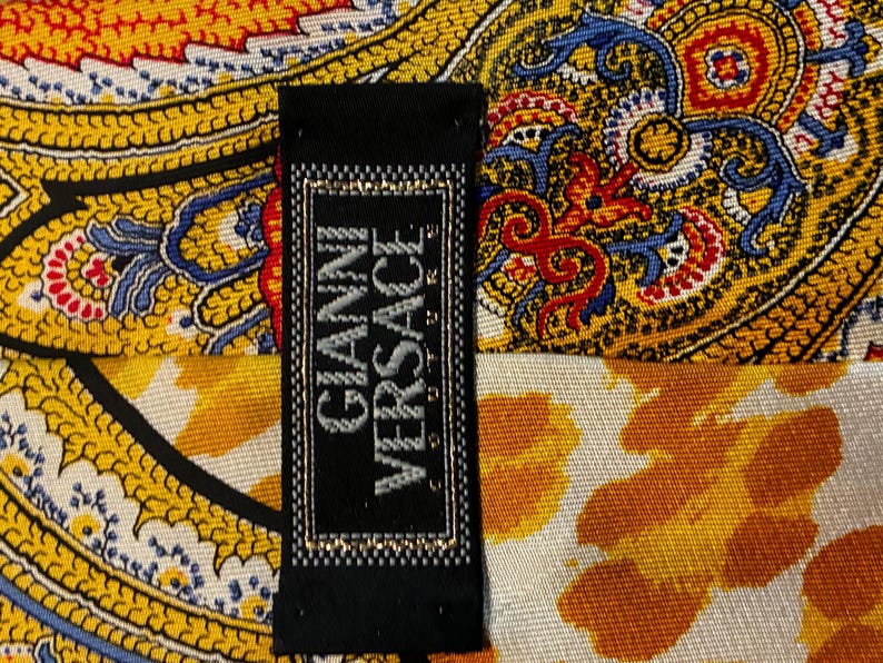 May include: A black label with the text "GIANNI VERSACE" in silver lettering is displayed against a backdrop of a colorful patterned fabric. The fabric features a paisley design with yellow, orange, blue, and red hues.