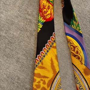 May include: A black silk scarf with ornate designs. One section features a yellow and red face with green accents. Another section has a tiger print pattern in yellow and brown. The scarf has a detailed border with gold and blue accents.