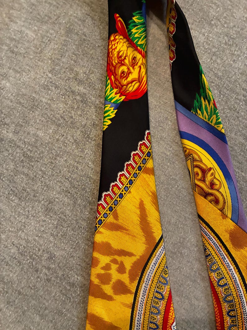 May include: A black silk scarf with ornate designs. One section features a yellow and red face with green accents. Another section has a tiger print pattern in yellow and brown. The scarf has a detailed border with gold and blue accents.