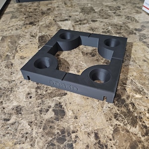 May include: A dark gray, square-shaped 3D-printed object with four circular openings and the text "QOB3D" on the side. The object is designed with interlocking sections and sits on a marble-patterned surface.