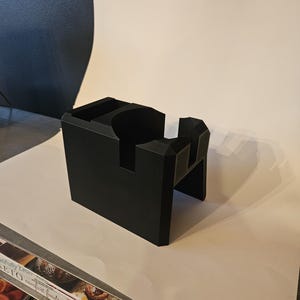 以下が含まれることがあります： 黒色の幾何学的なコーヒーアクセサリー。長方形のベースと切り欠きがあり、ノックボックスまたはポルタフィルターホルダーの可能性があります。マットブラックの素材でできており、白い表面に置かれています。