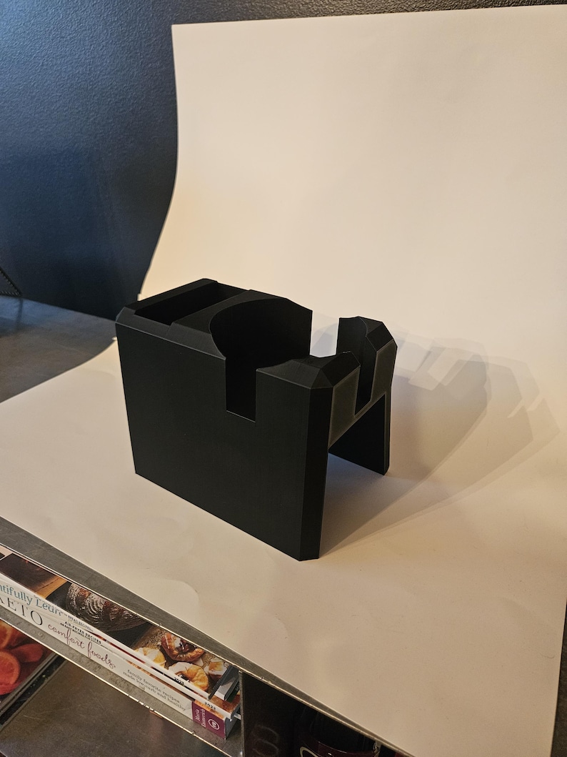 以下が含まれることがあります： 黒色の幾何学的なコーヒーアクセサリー。長方形のベースと切り欠きがあり、ノックボックスまたはポルタフィルターホルダーの可能性があります。マットブラックの素材でできており、白い表面に置かれています。