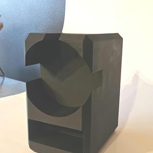 以下が含まれることがあります： 暗い灰色の幾何学的な彫刻で、立方体の基部があります。彫刻には、大きな円と長方形のスロットを含む、切り抜かれた形状があります。デザインはモダンで抽象的で、室内装飾に適しています。