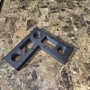 May include: A dark gray, L-shaped bracket with two circular holes and rectangular cutouts. The bracket is made of a solid material and appears to be designed for structural support or joining components. The item is shown on a brown and tan patterned surface.