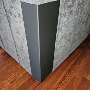 May include: Close-up of a gray upholstered sofa corner with a dark gray metal corner accent. The sofa sits on a wooden floor with a reddish-brown finish. The metal accent is a dark gray color.