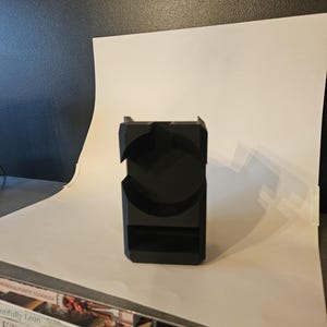 以下が含まれることがあります： 黒色の幾何学的な電話スタンド。曲線的な切り欠きと長方形のベースがあります。スタンドはマットな素材でできており、電話を立てておくように設計されています。デザインはモダンでミニマリストです。