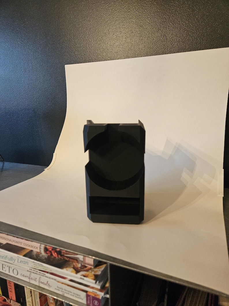 以下が含まれることがあります： 黒色の幾何学的な電話スタンド。曲線的な切り欠きと長方形のベースがあります。スタンドはマットな素材でできており、電話を立てておくように設計されています。デザインはモダンでミニマリストです。