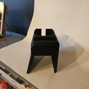 以下が含まれることがあります： 長方形のベースと角度の付いた脚を備えた、黒色の幾何学的なスタンド。上部には垂直のスロットと水平の棚があります。スタンドは無地のマットな素材でできており、アイテムを保持するように設計されています。