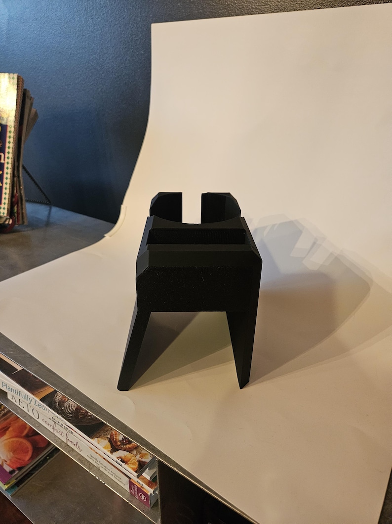 以下が含まれることがあります： 長方形のベースと角度の付いた脚を備えた、黒色の幾何学的なスタンド。上部には垂直のスロットと水平の棚があります。スタンドは無地のマットな素材でできており、アイテムを保持するように設計されています。