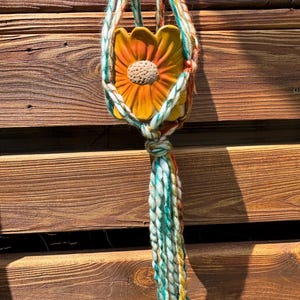 May include: A yellow and orange ceramic flower pot holder suspended by a macrame plant hanger. The macrame is made of white, teal, orange, and yellow yarn. The flower pot has a textured center.
