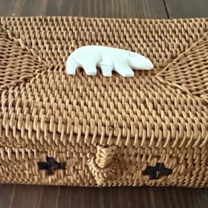 May include: Woven rectangular box with a light brown finish. A white polar bear figurine adorns the lid. The box features a woven clasp and decorative black accents. The box is made of woven rattan.