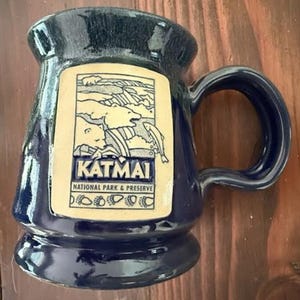 May include: A navy blue ceramic mug with a cream-colored rectangular graphic featuring a landscape scene and the word "KATMAI" in bold letters. The mug has a rounded handle and a wide base.