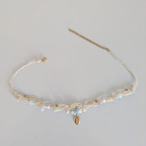 Peut inclure: Un collier en macramé crème avec des perles bleues clair et blanches, et un pendentif coquillage doré. Le collier a une chaîne et un fermoir dorés.