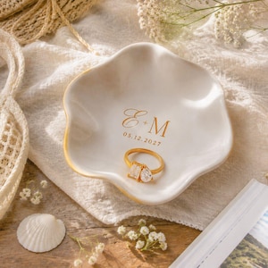 May include: A white ceramic trinket dish with wavy edges featuring gold lettering that reads E + M and the date 05.12.2027. A gold ring with a pear-shaped and a rectangular-cut clear stone rests in the centre of the dish, which has a gold rim.