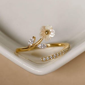 May include: A gold ring with a floral design. The ring features a small, white flower with a gold centre and two leaf-shaped accents. The band is adorned with small, clear gemstones. The ring is set against a white background.