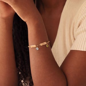 May include: Gold-toned bracelet with rectangular nameplates and dangling charms. The bracelet features a delicate chain and is adorned with a small blue gemstone and a green charm. The bracelet is worn on a person's wrist.