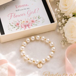 May include: A white pearl bead bracelet with small gold-tone metal spacers and a round gold-tone charm featuring the letter E. The bracelet is presented in an open cream-colored box with a card that reads, Emma, Will you be my Flower Girl?