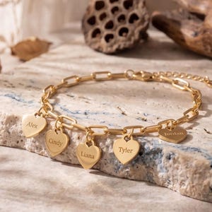 May include: A gold-colored bracelet with a chain link design. The bracelet features heart-shaped charms engraved with names: Alex, Cindy, Liana, Tyler, and Jordan. The bracelet is displayed on a textured stone surface.