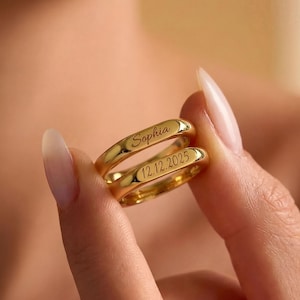 May include: Two gold wedding bands, one stacked on top of the other, are held between fingers. The top band is engraved with the name "Sophia", and the bottom band has the date "12.12.2025". The rings have a polished finish and a rounded shape.
