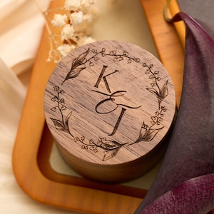 May include: A round wooden ring box with a floral wreath design and the initials "K & J" engraved on the lid. The box is made of a light brown wood and sits on a wooden tray. A dark purple calla lily is in the background.