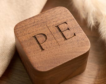 May include: A square wooden ring box with the initials "P E" engraved on the lid. The box is made of a dark brown wood and sits on a wooden tray. The background includes a beige cloth and dried ornamental grass.