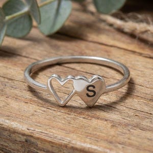 May include: A silver ring with two heart designs. One heart is open, and the other has the letter "S" in black. The ring is displayed on a wooden surface, with green leaves visible in the background.