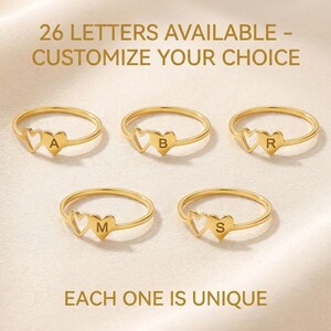 Peut inclure: Des bagues dorées avec des motifs en forme de cœur et des lettres individuelles. Les bagues sont présentées sur un fond doux et neutre. Le texte indique : "26 LETTERS AVAILABLE - CUSTOMIZE YOUR CHOICE" et "EACH ONE IS UNIQUE".