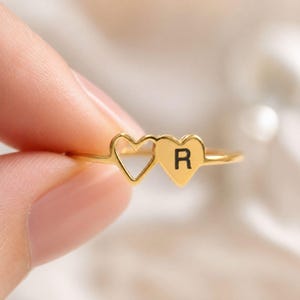 Peut inclure: Une bague dorée ornée de deux cœurs. L'un est plein avec la lettre "R" en noir, l'autre est ajouré. La bague est tenue entre les doigts, mettant en valeur son design délicat et romantique.