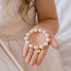 May include: A stretch bracelet featuring alternating white faux pearl beads and small gold-toned metallic spheres. A circular gold-toned charm hangs from the bracelet, engraved with the capital letter S in a simple, clean sans-serif font.