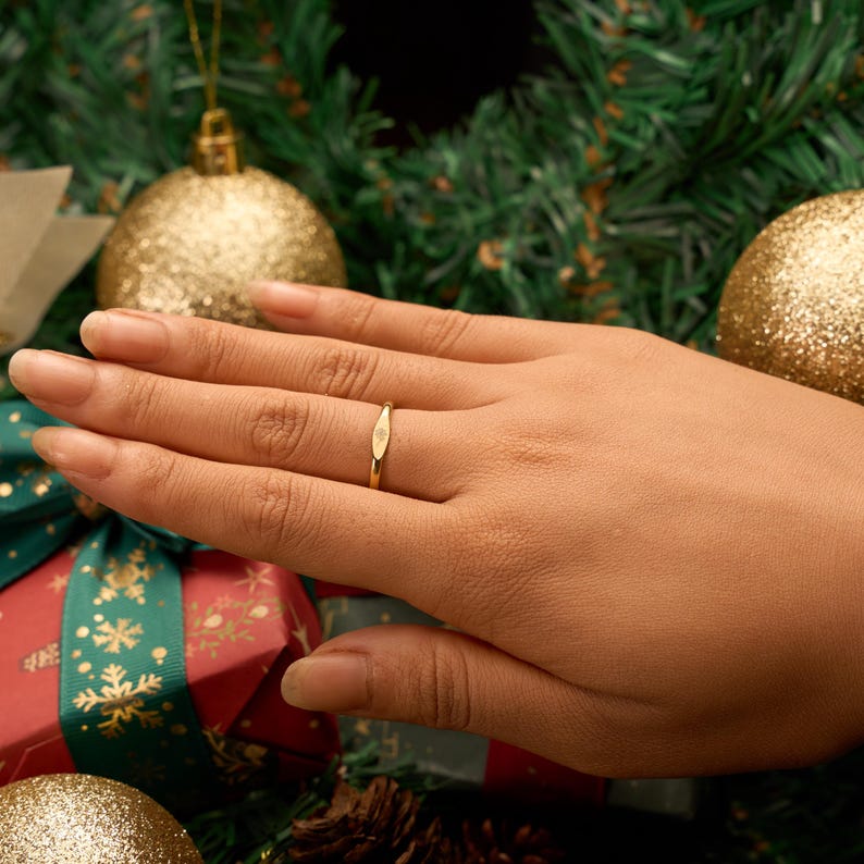 May include: A gold ring with a small, oval-shaped design is displayed on a hand. The ring is the focal point, set against a festive backdrop of Christmas ornaments and a wrapped gift. The ring's simple elegance is highlighted by the warm lighting.