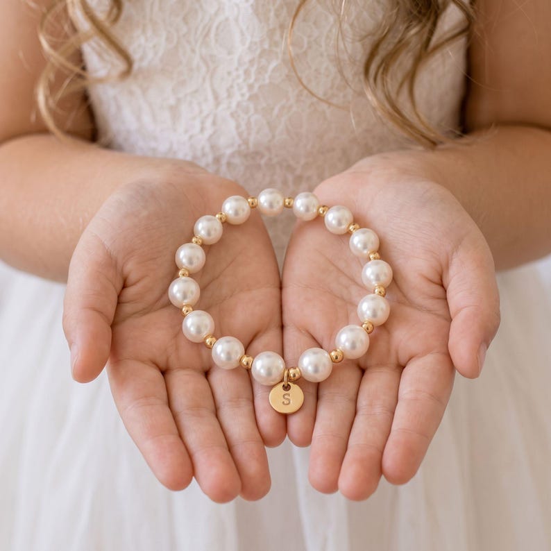 May include: A beaded bracelet featuring alternating white pearls and small gold-toned spherical spacers. A circular gold-toned charm hangs from the bracelet, engraved with the capital letter S in a simple, clean font, centered on the bottom.