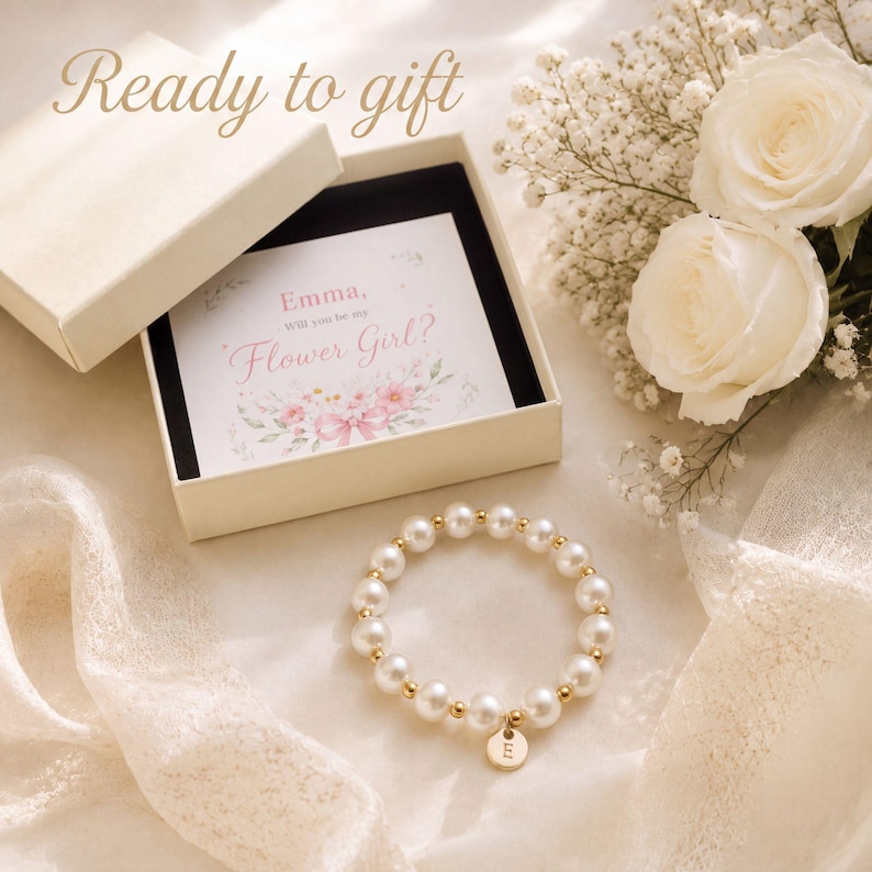 May include: A white gift box containing a card with the text Emma, Will you be my Flower Girl? and a floral illustration. Beside the box is a bracelet made of white pearls and gold beads with a round gold charm featuring the letter E.