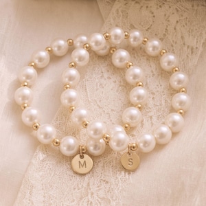 May include: Two stretch bracelets composed of round white faux pearls and small gold-toned metal spacer beads. Each bracelet features a single circular gold-toned metal charm, one engraved with the letter M and the other with the letter S.