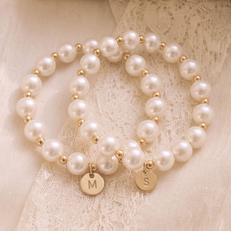 May include: Two stretch bracelets composed of round white faux pearls and small gold-toned metal spacer beads. Each bracelet features a single circular gold-toned metal charm, one engraved with the letter M and the other with the letter S.