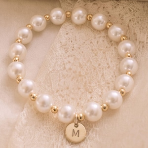 May include: A stretch bracelet featuring alternating white faux pearl beads and small gold-toned metal spacer beads. A circular gold-toned metal charm hangs from the bracelet, engraved with the capital letter M in a simple sans-serif font.
