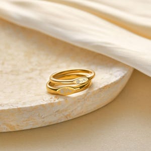 以下が含まれることがあります： 2つのゴールドカラーのリングが重ねられています。各リングは楕円形で、前面に花の模様が刻まれています。リングは明るい色のテクスチャのある表面に置かれています。